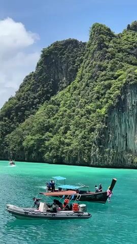 The video shows a scenic view of a body of water with several boats. In the foreground, a gray inflatable boat with people aboard is visible. To its right, a long-tail boat with people is also present. In the background, a larger boat with a canopy and people is moving. Further in the distance, another boat can be seen. Towering, lush green cliffs rise majestically behind the boats, creating a dramatic backdrop. The water is a vibrant turquoise color, indicating clear and shallow conditions. The sky is partly cloudy with patches of blue.