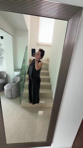 A woman with short brown hair, wearing a gray sleeveless top and matching pants, takes a selfie in a large mirror. She is standing on the bottom step of a staircase in a modern home with white walls and light tile flooring. The mirror reflects part of a living room with gray furniture, a glass railing, and abstract art on the wall. The staircase leads to a window with sunlight streaming through. The woman holds her black phone up to her face, posing with rings and bracelets on her left hand. She appears to be indoors during the day.
