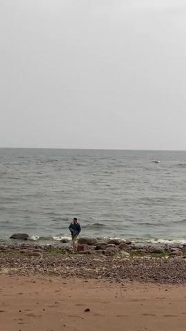 A person walks along a rocky shoreline next to the water. The person is wearing a blue jacket and khaki shorts. The sky is overcast and gray. The water is choppy. The shoreline is covered in rocks and some sand.