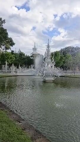 The video captures a wide shot of a serene outdoor scene featuring a pond with a fountain. The fountain, a centerpiece of the water feature, is intricately designed with what appears to be white, ornate sculptures, possibly in a mythical or artistic style. Water sprays upwards from the fountain, creating a dynamic element against the tranquil water surface. In the background, a picturesque landscape unfolds. Lush green trees form a natural backdrop, framing the scene. Beyond the trees, a striking white building or structure with intricate architectural details stands tall, possibly a temple or a significant monument, adding to the aesthetic appeal. The sky above is a mix of white clouds and patches of blue, suggesting a pleasant day. The overall ambiance is one of peacefulness and visual beauty, with the interplay of water, architecture, and nature creating a captivating spectacle.