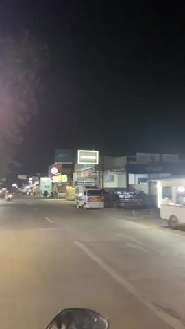 The video shows a nighttime street scene from the perspective of someone riding a motorcycle. The street is paved and illuminated by streetlights and the neon signs of various businesses. On the right side of the street, there are shops with prominent signs for "Oppo" and "Samsung," along with other businesses displaying colorful advertisements. A white van with blue and red accents is parked on the side of the road. Several food stalls or small shops with green and yellow facades are also visible. On the left side of the street, there are more shops and signs, including a circular red and white sign. People are seen on motorcycles and standing near the shops. A tree with dark foliage is visible on the far left, partially obscuring the sky. The overall atmosphere is that of a busy commercial area at night.