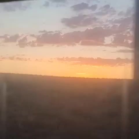 The video captures a view of a sunset or sunrise through a window. The sky is a blend of soft orange, pink, and pastel blue hues, with scattered clouds. The sun is visible near the horizon, casting a warm glow. Below the sky is a hazy, muted landscape, possibly a field or plain, with a subtle gradient of browns and grays, suggesting distance and atmospheric conditions like dust or fog. The light creates a soft, diffused effect across the scene. The video has a slight blur and some light flares are visible, indicating it was likely recorded from a moving vehicle or through glass.