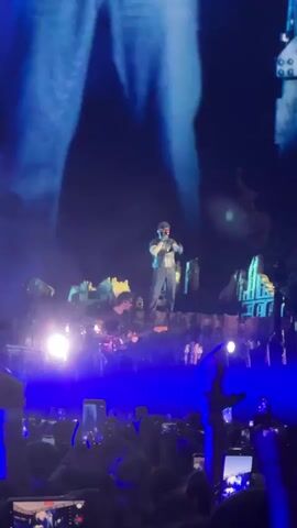 A concert stage at night is depicted. A male singer, wearing a black outfit with a vest and a mask covering his face with red glowing eyes, stands on a raised platform. He is holding a microphone and gesturing with his hands. To his left, a guitarist, wearing glasses and a black shirt, plays a red electric guitar. The stage is decorated with structures resembling buildings and is lit by spotlights. A large blue backdrop is visible behind the performers. In the foreground, the audience is holding up numerous mobile phones, most of which are recording or taking pictures. The overall atmosphere is dark and energetic.
