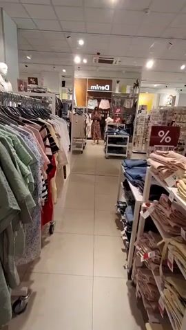 A video shows the interior of a clothing store. The camera is positioned between racks of clothing, looking down an aisle. In the distance, a woman is visible in a mirror, wearing a floral dress and looking at herself. She appears to be in the process of examining or trying on clothing. Various clothing items, including shirts, jeans, and dresses, are displayed on racks and shelves throughout the store. Discount signs with a percentage symbol are visible in some areas. The store appears to be well-lit with fluorescent lighting.