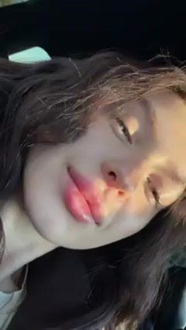 The video shows a close-up of a young woman's face. She has brown hair and pink lips. She is looking directly at the camera. She is inside a car. There is sunlight on her face.