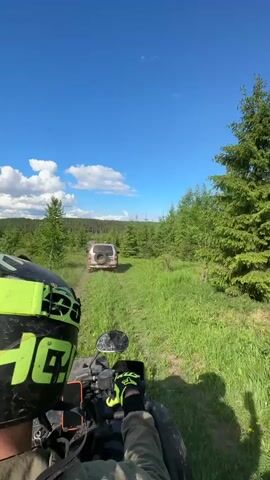 A first-person perspective video shows someone riding an ATV on a grassy trail towards a silver SUV in front. The ATV rider wears a black and neon green helmet, gloves, and tan sleeves. A smartphone is visible on the ATV handlebars. The SUV is driving away on the dirt track, with a spare tire mounted on its back. The background features a forest with evergreen trees and a bright blue sky with scattered white clouds. The environment is sunny and appears to be a rural or wilderness setting.