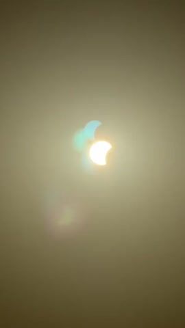This is a video of a solar eclipse, captured in a hazy or foggy environment. The sun is partially obscured, creating a crescent shape in the sky. There appear to be multiple overlapping images of the eclipsed sun, suggesting either a deliberate photographic effect or atmospheric conditions causing light refractions. The overall color palette is muted, with the sun's brightness standing out against the diffused background.