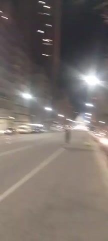 The video begins with a blurry shot of a street at night, illuminated by streetlights and car headlights. The camera pans to the right, showing parked cars along the side of the road and the facade of a building. In the second clip, a person wearing a red jacket and a cap is seen waving a large flag with blue and white stripes, possibly the flag of Argentina, while walking on the street. The background shows a tall building and streetlights. The video then briefly shows a blurry scene of people walking away from the camera in what appears to be a courtyard or walkway. The final clip is a fast-moving, blurry shot of a street at night with cars passing by, some with glowing lights.