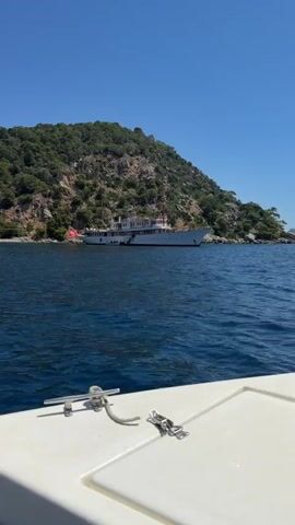 The video captures a serene daytime scene on the clear blue waters of Poros, Greece. The perspective is from the bow of a small, white boat, with its metal cleats and a rope visible in the foreground.

The primary subject is a large, classic-style white yacht, anchored a short distance from a lush, green, and rocky coastline. The yacht features multiple decks, numerous portholes along its hull, and what appears to be a gangway or steps extending from its side towards the water. A red flag is visible on a mast near the bow of the yacht.

In the background, a steep hill covered with dense green foliage and exposed rocky patches rises from the water's edge under a brilliant clear blue sky. The water itself is a deep blue, showing gentle ripples on its surface. The overall impression is one of a calm, sunny day in a picturesque coastal setting. No people or visible text are discernible in the video.