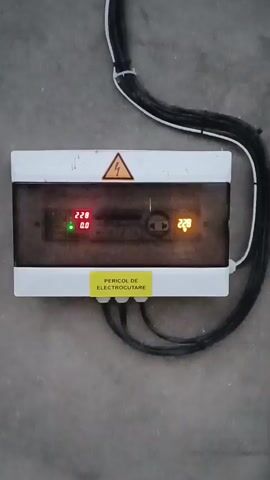 The video shows a close-up, slightly shaky, eye-level shot of a white electrical box mounted on a gray concrete wall. The box has a clear plastic cover, revealing two digital displays. The left display shows "22.8" in red and "0.0" in green. The right display shows "22.7" in orange. Above the displays, there is a yellow triangle with a black lightning bolt, a standard symbol for high voltage. A yellow sticker with black text that appears to read "PERICOL DE ELECTROCUTARE" is affixed to the bottom of the box, which translates to "DANGER OF ELECTROCUTION". Three thick black cables exit the bottom of the box, and several more black cables are bundled together and run vertically along the top right of the box. The lighting is dim, and the focus is on the electrical box and its displays, which fluctuate slightly throughout the short clip.