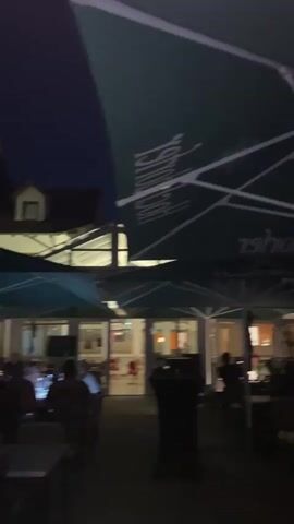 The video shows an outdoor seating area of a restaurant or bar at night. Large green umbrellas with white lettering are prominently displayed. Tables and chairs are set up on a patio. Several people are seated at the tables, some are looking at their phones, others are engaged in conversation. The building behind the patio is illuminated. The atmosphere appears relaxed.