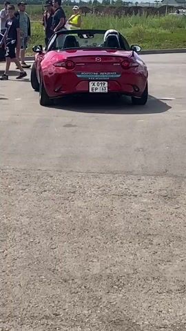 The video shows a red Mazda MX-5 convertible parked on a paved surface. There are three people standing to the left of the car, looking towards it. The car has its top down and a person wearing a helmet is sitting in the driver's seat. The license plate of the car is visible and reads "X 019 EP 63". In the background, there is a grassy area with some trees and houses. The sky is cloudy. The car appears to be stationary for the entire duration of the clip.