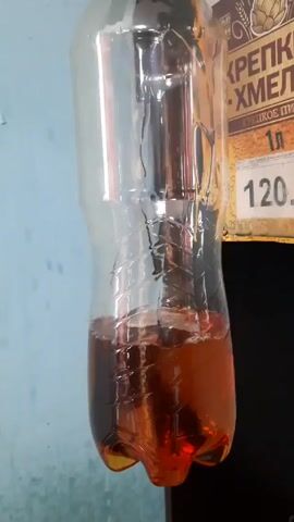 A clear plastic bottle hangs suspended from a black fixture.  Inside the bottle, about a third full, is a brown liquid. The liquid is agitated, creating ripples and swirling patterns within the bottle.  A price tag is partially visible to the right of the bottle, displaying the text "КРЕПКИ ХМЕЛИ • ХМЕЛИ" and the price "120". The background consists of a light blue wall.