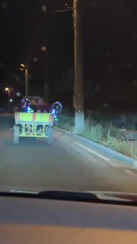 The video shows a rear view of a flatbed trailer being pulled at night. The trailer is yellow with red stripes and decorated with colorful LED lights and a large clear balloon. Two people are visible in the trailer, one wearing a red shirt. The trailer appears to be moving along a paved road with a curb and sidewalk. The camera is inside a vehicle, and the dashboard is visible in the foreground. The background is dark, with some streetlights and trees. The scene suggests a festive or celebratory atmosphere.
