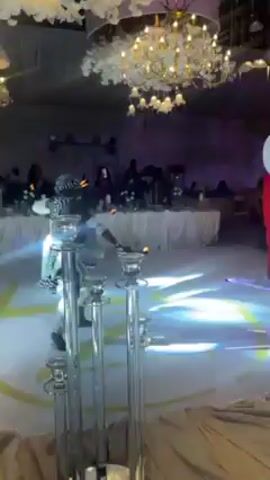 A person wearing a red suit, hat, and white face covering, plays a percussion instrument. They are in what appears to be a wedding reception hall with tables, chairs, and other guests. The hall has elaborate chandeliers and lighting. The person is walking around the hall while playing. Another person in a pink dress stands nearby.