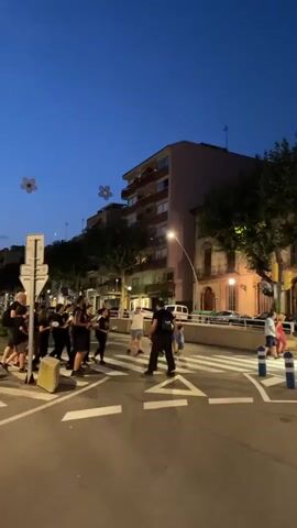 This video captures a vibrant street scene in Arenys De Mar, Spain, as dusk settles. A group of people, many dressed in dark clothing, are gathered in a pedestrian crossing. The primary activity appears to be a musical performance, with several individuals holding instruments, some of which resemble drums. The atmosphere is lively and engaging, with onlookers watching the performance.

The setting is urban, with apartment buildings lining the street. The sky is a deep blue, indicating either twilight or early evening. Decorative elements, like flower-shaped lights suspended across the street, suggest a festive occasion or a local event. Streetlights are also visible, casting a warm glow on the scene, and illuminating the white markings of the pedestrian crossing. The presence of a traffic light and street signs confirms the urban environment.

The people in the video are a mix of performers and spectators. The performers are actively engaged with their music, creating a sense of energy and rhythm. The spectators are observing the performance, some sitting on a concrete barrier, while others stand. The overall mood seems positive and communal. The time of day is clearly evening, given the blue sky and artificial lighting. The weather appears to be clear. There is no visible text in the video.