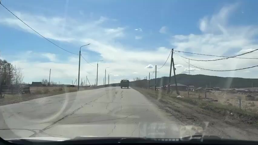 A view from inside a car driving on an asphalt road through a rural area. A truck is driving in the distance ahead of the car. There are utility poles along both sides of the road, with power lines spanning across the street. The road is cracked and shows signs of wear. On either side of the road, there is dry, grassy land, with scattered houses and wooden fences. In the background, a hill or low mountain range is visible under a blue sky with scattered clouds. The sun is shining brightly.