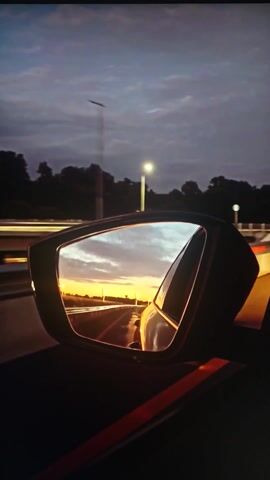 This media file is a short video clip, approximately 4 seconds long, captured from inside a vehicle. The primary focus is on the car's side-view mirror, which reflects a dramatic sunset. The sky in the reflection is a vibrant orange and yellow, with streaks of white clouds, indicating the sun is low on the horizon. The reflection also shows a road stretching into the distance, lined with trees and streetlights on the left side.

Outside the mirror's reflection, the sky is a deep twilight blue, with a few distant lights visible. The silhouette of trees forms a dark line against the horizon. To the left of the mirror, a directional sign with red and white chevron markings is briefly visible, suggesting the vehicle is on a road, possibly a highway or a main thoroughfare. The overall mood is serene and slightly melancholic, amplified by the fading light of day and the solitary perspective from within the car. The time of day is clearly sunset or dusk. The location is implied to be Belorechensk, Russia, based on the provided context, though no specific Russian landmarks are identifiable within the frame. There is no visible text in the media file.