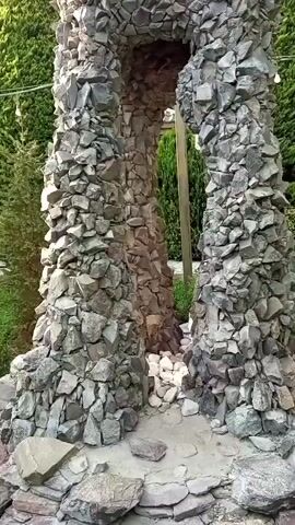 This video captures a picturesque garden setting in Buzovna, Azerbaijan. The scene is dominated by lush greenery, featuring a variety of trees and meticulously trimmed bushes. Prominently displayed is an interesting stone archway, constructed from rough-hewn gray and brown stones, adding a rustic and natural element to the landscaping.

In the background, a wooden gazebo with a tiled roof suggests a place for relaxation or gatherings. A wooden bench with a metal frame is also visible, inviting visitors to sit and enjoy the surroundings. The pathway leading through the garden is paved with decorative tiles arranged in a striking zigzag pattern of gray, white, and pinkish-red hues. String lights are suspended between trees, hinting at the possibility of evening enjoyment in the garden.

The time of day appears to be late afternoon or early evening, with soft, diffused light illuminating the scene. The weather seems pleasant, with no indications of rain or strong winds, contributing to the tranquil atmosphere. The overall impression is one of a well-maintained and aesthetically pleasing outdoor space, likely a private garden or a public park designed for leisurely enjoyment. There is no visible text in the video.