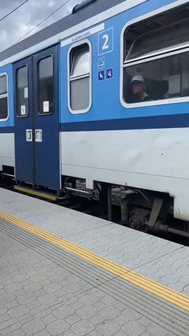A blue and white train is stopped at a platform. The train cars are connected by blue doors. The train has windows, and people can be seen inside. The platform is made of gray tiles with a yellow tactile warning strip along the edge. The train has the České dráhy logo on it. The sky is cloudy.