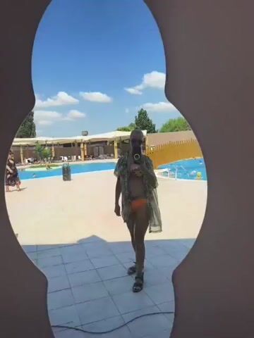 A person stands in front of a mirror, taking a selfie. The person is wearing a bikini and a sheer cover-up. In the background, there is a swimming pool with people in it, lounge chairs, and trees. The sky is blue with white clouds. The mirror is framed in a cat-like shape.