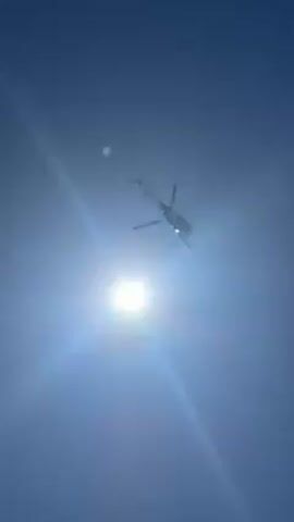 A helicopter flies over a sunny beach. The camera is pointed upwards, first showing the helicopter against the bright blue sky and then showing the helicopter above the beach, crowded with people, umbrellas, and tents.