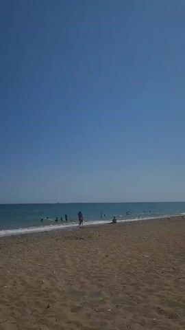 A static medium shot captures a wide view of a sandy and pebbled beach meeting the turquoise ocean under a clear blue sky. Small waves are visible breaking on the shore. Several people are present; two adults stand near the waterline, one wearing a red top. Other individuals are seen wading or swimming in the ocean. One person is seated on the sand to the right. The scene is brightly lit, indicative of a sunny day.