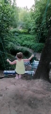 A young girl with blonde hair pulled into a ponytail, wearing a yellow sleeveless top and denim shorts, is swinging on a blue and white swing suspended by green ropes from a tree. She is positioned in a wooded area with dense green foliage on either side. The ground in the foreground is dirt, and there appears to be a small stream or ditch with debris below the swing. The girl is facing away from the camera, with her arms extended to the sides. The camera is angled slightly upwards, capturing the girl and the trees.