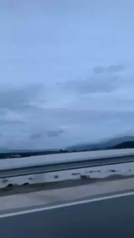 The video depicts a view from inside a moving car, driving along a highway. The sky is overcast with grey clouds. In the distance, a body of water is visible, with land and mountains beyond it. The car's side mirror is visible in the bottom left of the frame, offering a glimpse of the passing landscape. The focus remains on the scenery outside the car window as it moves along the road.