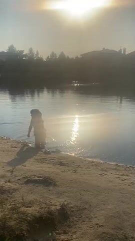 A lone child, partially submerged in water, stands at the edge of a sandy shore. The sun, positioned high in the sky, casts a brilliant glare that reflects off the water's surface, creating a shimmering effect. The child's silhouette is visible against the bright light. The environment appears to be a natural setting, possibly a lake or river, with trees and buildings visible in the background. The overall mood is serene, bathed in the warm glow of the setting sun.