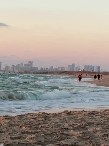 The video shows a beach scene at sunset. The sky is a pale pink and orange, with a few clouds. The ocean is a vibrant green, with waves breaking on the shore. In the distance, a city skyline is visible. There are several people walking along the beach. In the foreground, the sand is visible, with ripples from the wind. The video appears to be shot from a low angle, looking out towards the ocean.