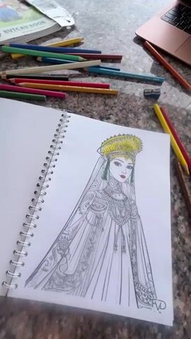The video pans across a table with art supplies and a laptop. In the foreground, a notebook lies open to a drawing of a woman in traditional clothing, with a yellow headdress and intricate detailing on her dress. Colored pencils are scattered around the notebook, along with a pencil sharpener. To the right, a pink laptop is open, displaying a webpage. In the background, a landscape with rolling hills and a building is visible under a cloudy sky. The table appears to be made of granite.