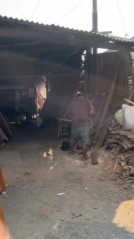 The video shows a man in a brown shirt and shorts operating a circular saw to cut wood. He is standing barefoot in a dusty outdoor area with a cluttered environment. To his left, there are stacks of lumber and what appears to be a workshop or storage area with a corrugated metal roof. To his right, there are large piles of scrap wood, bricks, and a large white bag. The ground is dirt and debris. Another person is partially visible in the background within the workshop area. The lighting suggests it is daytime with sunlight.
