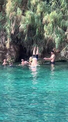 A group of young men are swimming and jumping into a clear, turquoise body of water. The water is surrounded by lush green reeds and rocky cliffs. One man is captured mid-air as he jumps into the water, creating a large splash. Another man is reaching out to him from the water. The other men are swimming and observing the scene. The sunlight is reflecting off the water, creating a beautiful and inviting atmosphere.