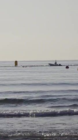 A wide shot captures a calm ocean scene under a hazy sky. In the distance, a small boat with an outboard motor navigates the water. A yellow buoy stands tall near the left side of the frame, and a red buoy floats closer to the right. Gentle waves lap at the shore in the foreground, with the edge of a sandy beach visible at the very bottom. The sun appears to be low in the sky, casting a soft light.