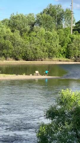A person wearing a blue shirt and black shorts stands on a small sandbar in a river. They are facing the water, with trees and a power line visible in the background. A chair and what appears to be a log or stump are also on the sandbar. The river has a gentle current. The sky is blue with some clouds.