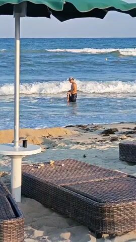 The video shows a man wading in the ocean. He is standing in the water, which is up to his waist. He appears to be holding something in his hands. The beach is sandy, and there are lounge chairs and an umbrella in the foreground. The ocean is blue and there are small waves. The sky is clear.
