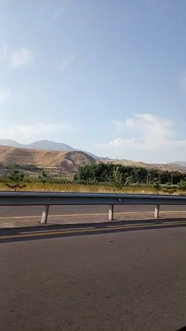 The video captures a roadside view from a moving vehicle, likely a car, on a highway. The sky is clear blue with a few scattered white clouds. Rolling hills with dry, tan-colored vegetation form the background, interspersed with patches of green trees. A metal guardrail with concrete posts runs along the side of the road, separating the highway from the landscape. Yellow lane markings are visible on the asphalt road surface. A black SUV passes by in the opposite direction, followed by a white van with tinted windows. The scene suggests a rural or semi-rural highway setting on a sunny day.