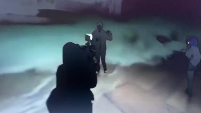 The video displays a first-person perspective of a dark-colored handgun being fired. In the foreground, the handgun is visible, and in the background, a person is seen falling backward, seemingly recoiling from a shot. Smoke or mist is present around the falling individual, obscuring details. The environment appears to be outdoors with some structures visible in the distance. The footage is blurry and appears to be taken at night or in low light conditions.