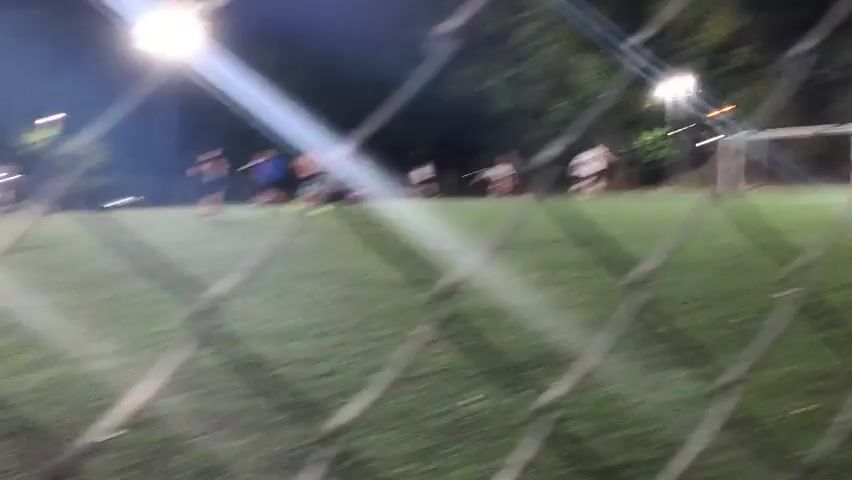 Through a chain-link fence, a soccer game unfolds on a green turf field at night, illuminated by bright stadium lights. Several players are visible, moving across the field. Behind the field, trees form a dark backdrop.