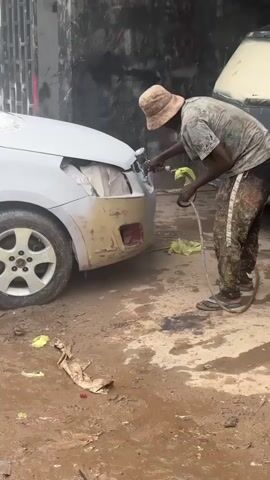 A person wearing a bucket hat, t-shirt, and paint-splattered pants is spray-painting the front of a light-colored car in what appears to be an outdoor repair shop or garage. The car's front bumper is partially primed, and its headlights are covered.  The person is using a spray gun connected to a hose. The ground is muddy and littered with scraps of plastic and debris.  In the background, a textured wall with graffiti or paint splatters can be seen, along with a partially covered vehicle.
