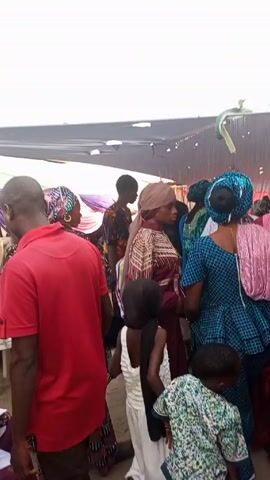 The video shows a gathering of people under a temporary structure that appears to be a tent or awning. The majority of the individuals are dressed in traditional African attire, particularly women in vibrant, patterned dresses and headwraps. The setting seems to be an outdoor event, possibly a celebration or social gathering. People are standing and moving around, suggesting they might be conversing or waiting for something to begin. The composition of the shot focuses on the crowd, providing a glimpse into the cultural context and the sense of community.