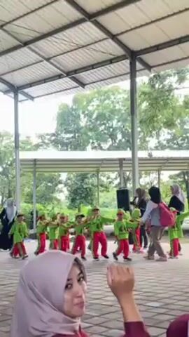 Here is a concise description of the video:

A group of young children, dressed in green shirts and red pants, are performing a dance routine under a covered outdoor area.  They are accompanied by several adult women wearing hijabs, who appear to be teachers or supervisors.  One of the women seems to be giving instructions with a microphone. In the foreground, a woman wearing a light purple hijab and a light-colored top looks towards the camera, smiles and waves. The setting appears to be a school or community gathering space with trees and greenery in the background.
