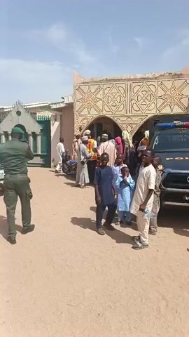 The video shows a street scene with a white pickup truck parked near a building with decorative Arabic script. A man in a green uniform walks towards the truck, while several people stand nearby. The scene then shifts to a band playing music on the street. Instruments like trumpets and drums are visible. A man in a white patterned shirt appears to be directing or dancing with the music. Several cars, including a blue one with "Police" written on it, are parked nearby. The environment appears to be a sunny day in a residential or public area. The buildings in the background have distinct architectural styles.
