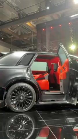 The video shows a black car on display at what appears to be an auto show. The car has custom wheels and red interior. The car doors are open, and its reflection is visible in the glossy black floor. The background includes the exhibition hall’s structural elements, stage lighting, and a decorative backdrop with red accents.