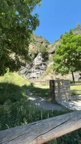 A video captures a cascading waterfall flowing down a rocky mountainside under a clear blue sky. Lush green trees frame the scene, with a dirt path leading towards the waterfall. In the foreground, a stone structure with a yellow warning sign is visible, along with a wooden fence. The camera zooms in on the waterfall, revealing its multiple tiers and the water's descent. A person in a light-colored shirt and shorts is seen climbing near the base of the waterfall. The video pans out to show the wider landscape, including a wooden walkway or bridge higher up on the mountain. The scene appears to be a natural park or hiking area.