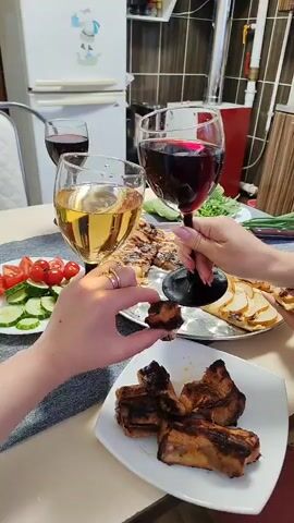 Two hands are visible on a light-colored table, one holding a clear wine glass with a light liquid and a small piece of grilled meat, while the other holds a dark-stemmed wine glass with red liquid. On the table, there is a white plate with several pieces of grilled meat, a metal tray with grilled potato slices and other food, and a white plate with sliced tomatoes and cucumbers. Green leafy vegetables and green onions are also present. In the background, a white refrigerator, a red appliance, and a tiled wall are visible.