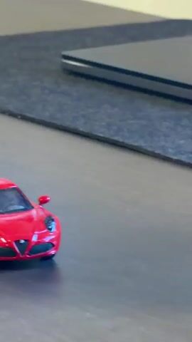 The video captures a close-up view of a small, red toy car, resembling a miniature sports car, resting on a dark, flat surface, likely a desk. In the background, a portion of a closed black laptop is visible, situated on a black desk mat. A blue cable is also partially seen extending into the frame from the right. The scene is set indoors, under ambient lighting conditions, without specific indicators for time of day or weather.

Initially, the red toy car is stationary. Around the 0:02 mark, a hand, only partially visible, enters the frame from the right side. The hand gently pushes the toy car, setting it in motion. The car then rolls smoothly across the dark surface towards the left, eventually moving out of the camera's view. The brief action depicts a simple interaction with the toy. No visible text is present on any of the objects in focus. The location context is Kafr Yasif, Israel.