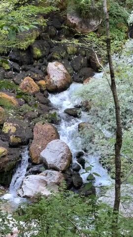 A slow-motion video shows a small waterfall cascading over mossy rocks in a forest. The water is clear and turbulent, creating white foam as it tumbles down. Large boulders, some covered in moss, are scattered around the waterfall, and lush green vegetation surrounds the scene. The overall atmosphere is serene and natural.