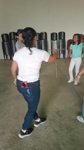 The video shows a group of people in a large room playing a game with a rope and plastic bottle. The group comprises mostly young women and girls. The game involves stepping over or ducking under the rope, which is held by two people. The video starts with a girl about to try to step over the rope without touching it. The room has a concrete floor and white walls with some red-brown trim. There are several rows of stacked black chairs in the background and some tables in another area of the room. The people appear to be having fun.